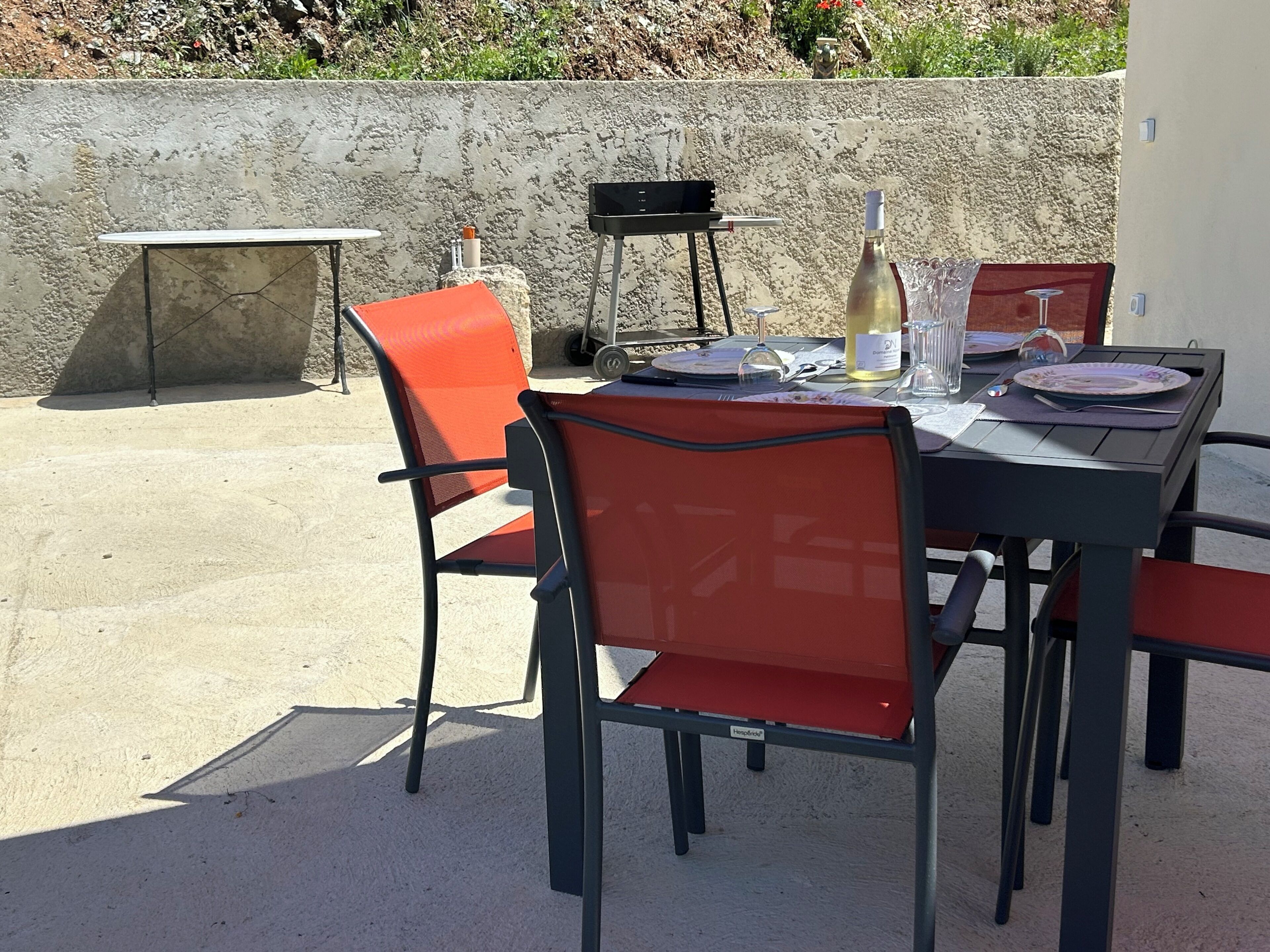 Un espace extérieur aménagé pour les repas, avec une table et des chaises, un barbecue et une bouteille de vin.