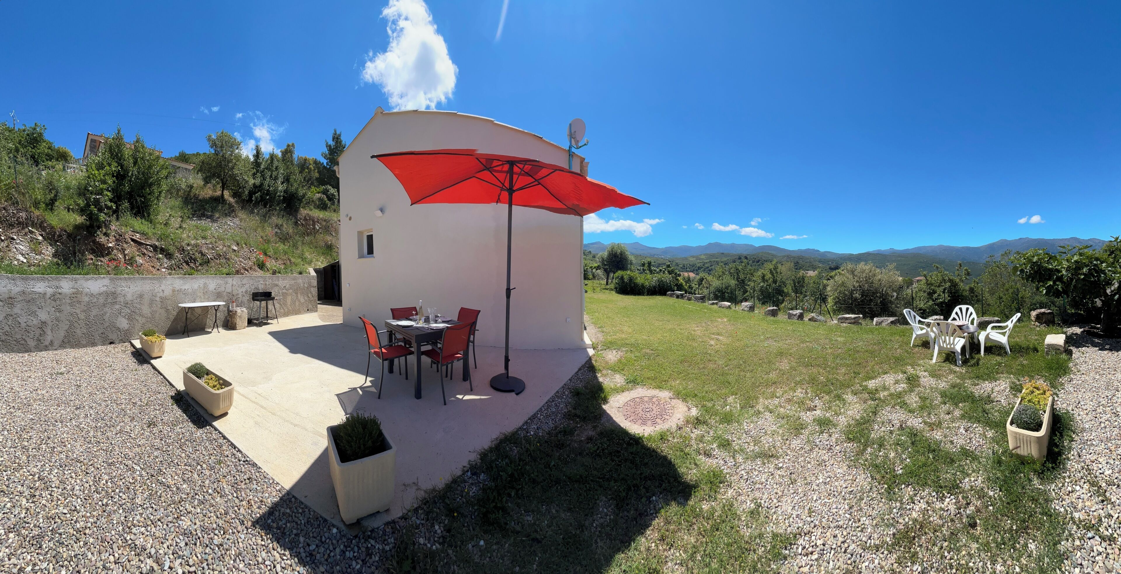 A small white building with a red umbrella, outdoor seating, and a view of mountains.