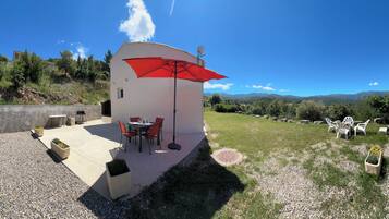 Un petit bâtiment blanc avec un parasol rouge, des tables à l’extérieur et une vue sur les montagnes.