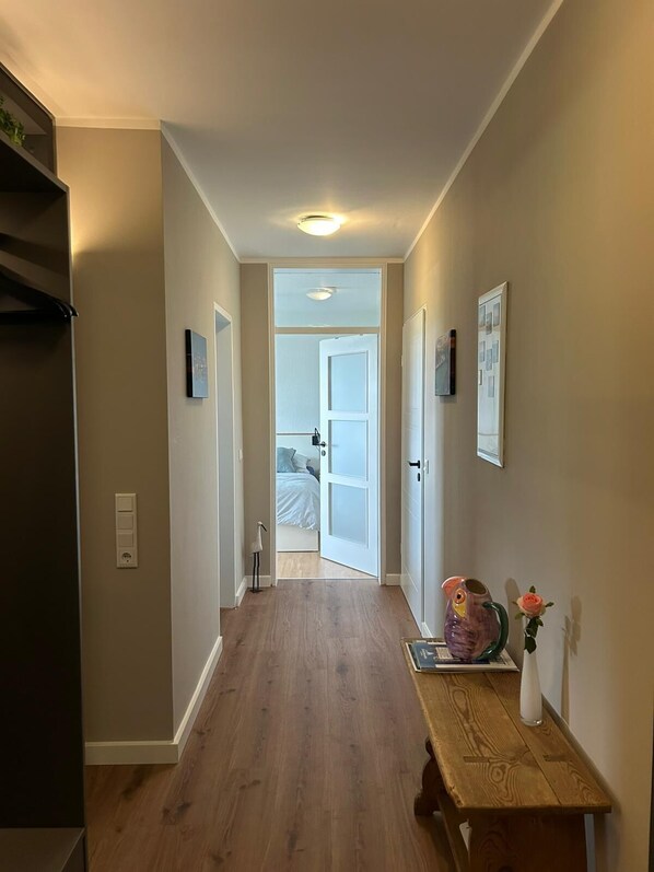En hall som leder till ett sovrum med en säng, ett träbord med en vas och en bok, och en dörr med en bilderram.