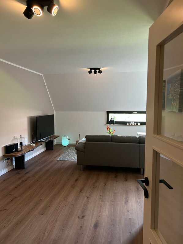 Una sala de estar moderna con un televisor de pantalla plana, piso de madera, un sofá gris y una cocina visible a través de una ventana.