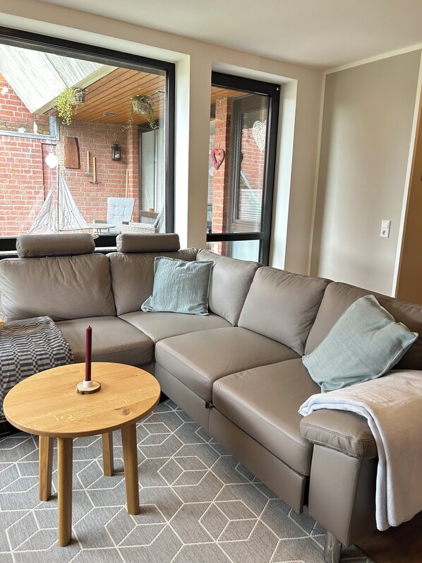 A modern living room with a sectional sofa, a round wooden table, and a view of a brick building through large windows.