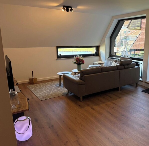 A modern living room with a sofa, a small table, a TV, and a window with a view of buildings. - Modern apartment with 61 m² and lake view in Bad Zwischenahn. (Bad Zwischenahn)