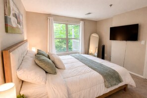 Quarto com uma cama grande, televisão fixada na parede e janela com cortinas.