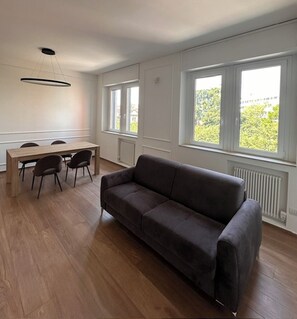 Una sala de estar moderna con un sofá, una mesa de comedor con sillas y ventanas grandes con vistas a la vegetación.