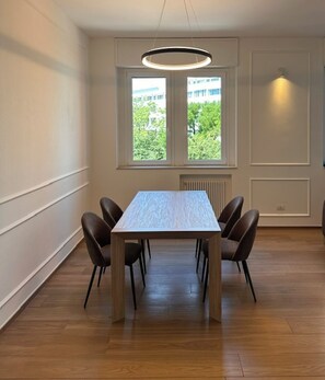 Un comedor con una mesa rectangular, seis sillas, una lámpara colgante y una ventana que ofrece vistas a zonas verdes y edificios.