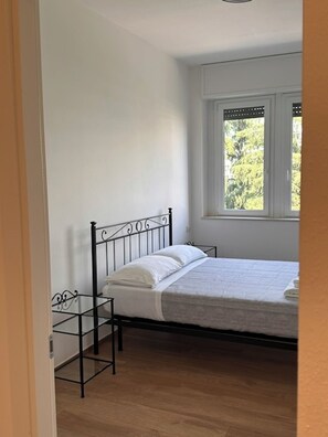 Un dormitorio con estructura de cama metálica, ventana con vista a árboles y suelo de madera.
