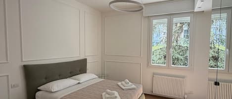 Une chambre à coucher comprenant un lit, une table de chevet, un luminaire suspendu, une fenêtre et un radiateur.