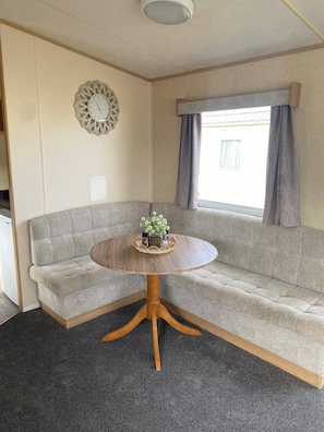 A compact living space with a sofa, a round wooden table, a decorative clock, and a window with curtains. - Cabin Sleeps 6 With Parking Near Beach (New Romney)