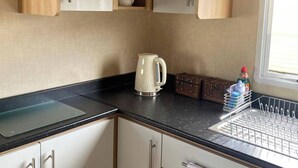 A compact kitchen with wooden cabinets, a kettle, a dish rack, and a window. - This Caravan is no Longer Available (New Romney)