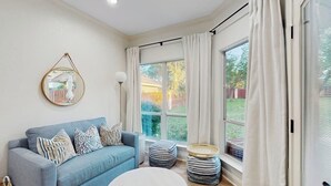 A living room with a sofa, ottomans, a round coffee table, a hanging mirror, and large windows with curtains. - Modern Home Near Lake LBJ With Enclosed Yard, Patio, Grill & Central AC (Horseshoe Bay)