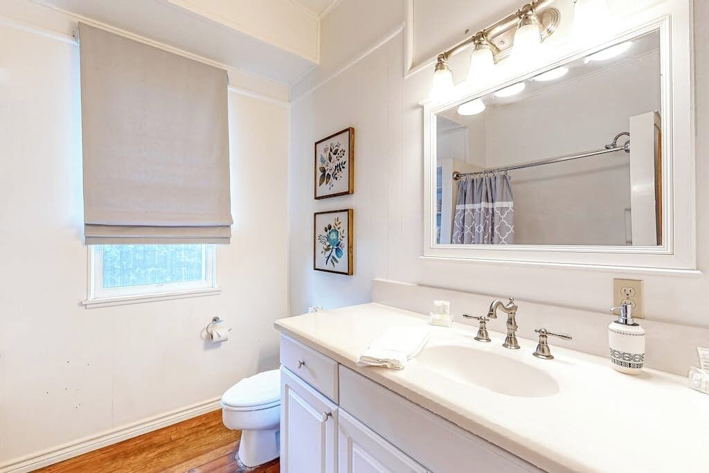 A bathroom with a white sink, vanity, and a large mirror. There is a window with a blind, and two framed pictures on the wall.
