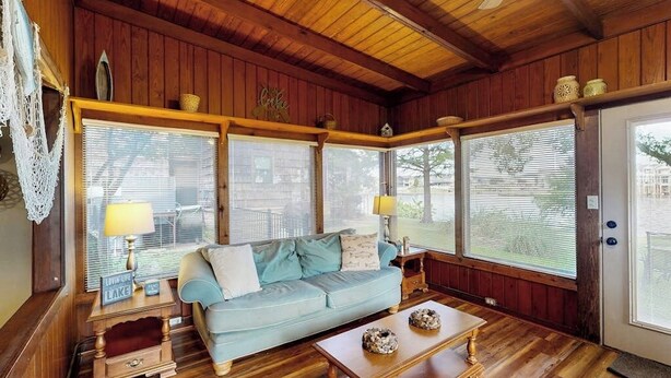 Ein gemütliches Blockhaus-Interieur mit Holzbalkendecke, einem hellblauen Sofa, zwei Nachttischen mit Lampen und großen Fenstern mit Jalousien.