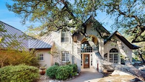A stone house with a central entrance, a covered porch, and a large tree in the foreground. - Sacred Vista Opulent Hill Country Gem Amazing Patio & Great View (Horseshoe Bay)