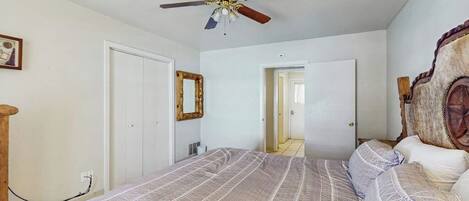 Une chambre à coucher avec un lit, un ventilateur de plafond, une tête de lit en bois et une porte donnant sur une autre pièce.