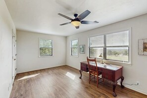 Une pièce dotée d’un plancher en bois franc, d’un ventilateur de plafond, d’un bureau avec une chaise et de grandes fenêtres équipées de stores.