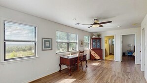 A spacious room with hardwood floors, a desk with a chair, a ceiling fan, and large windows with blinds. - Idyllic Home With Firepit, Gazebo, Patio, & Amazing Deck Views - Dog-friendly (Fredericksburg)