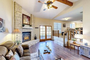 A spacious living area with a stone fireplace, a ceiling fan, and a view to the kitchen. - Golf Course-view Home With Yard, Grill, AC, & Washer/ Dryer - Dog-friendly (Leander)