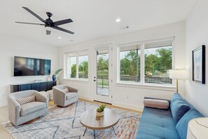 Una sala de estar moderna con ventilador de techo, televisor de pantalla plana, dos sofás, una mesa de centro y ventanas amplias con vista a follaje.