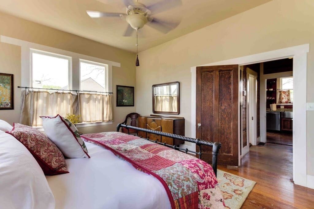 A bedroom with a bed, a desk, a chair, a ceiling fan, and a door leading to a kitchen.