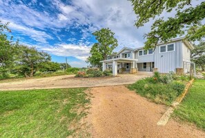 A two-story house with a covered porch, a gravel driveway, and surrounding greenery. - Historic Fredericksburg Farmhouse - Hill Country Views - Wine Trail - Snowbird (Fredericksburg)