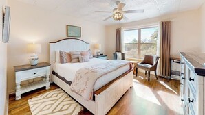 Un dormitorio con cama, mesitas de noche, una silla, cómoda, ventilador de techo y ventana con cortinas.