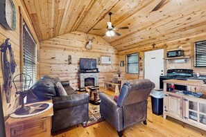 A log cabin interior with a fireplace, leather furniture, a ceiling fan, and a kitchen area. - Dog-friendly Gem on 11 Acres With Valley Views, Deck, Gas Grill, Fireplace (Fredericksburg)