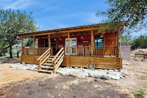 A rustic wooden cabin with a porch, a wheelbarrow, and a sign that reads 'RANCH.' - Dog-friendly Gem on 11 Acres With Valley Views, Deck, Gas Grill, Fireplace (Fredericksburg)