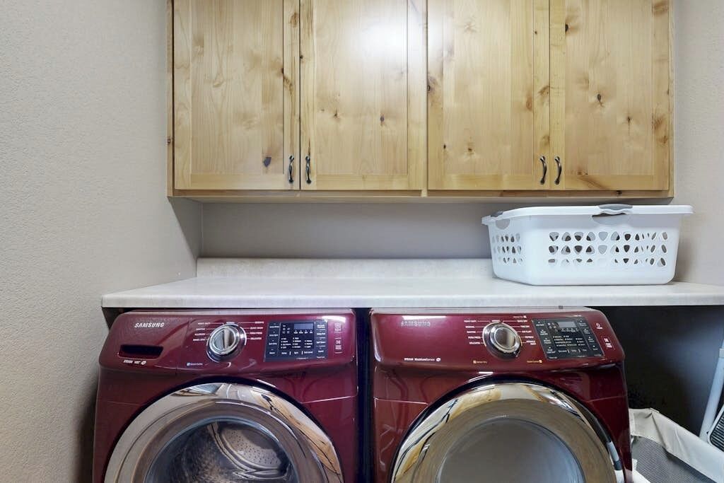 Two front-loading washing machines, a wooden cabinet above, and a white laundry basket on a countertop.