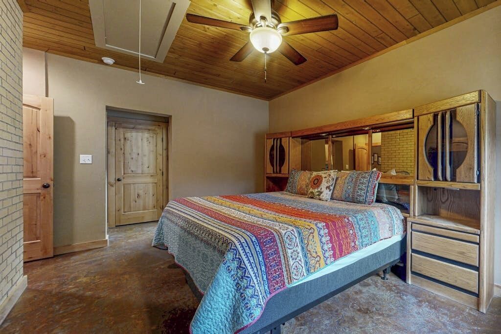A bedroom with a wooden ceiling, a bed with a colorful blanket, a wooden dresser, and a door.