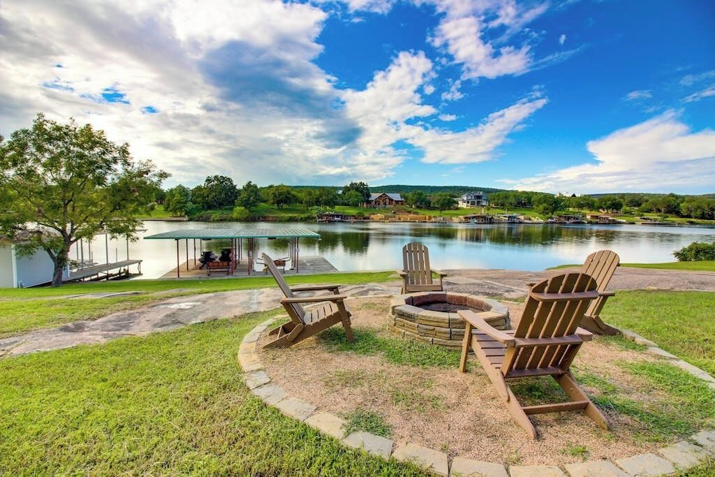 A lakeside patio with Adirondack chairs, a fire pit, and a view of a lake and distant buildings.