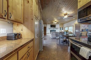 A kitchen with wooden cabinets, a granite countertop, a refrigerator, a microwave, and a dining area with chairs and a table. - Renovated Waterfront Home With Wifi, Lake Views, Dock & Boat Lift - Dogs OK (Burnet)