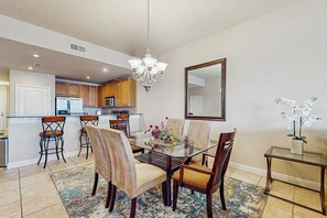 A dining area with a glass table, wooden chairs, a chandelier, and a kitchen in the background. - Dog-friendly & Spacious Condo With Lake Views, Large Balcony, & Pool (Horseshoe Bay)