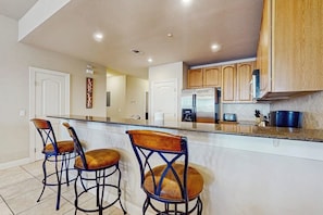 A kitchen with a central island, bar stools, wooden cabinets, and a refrigerator. - Dog-friendly & Spacious Condo With Lake Views, Large Balcony, & Pool (Horseshoe Bay)