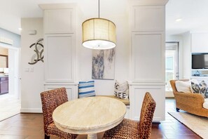 A dining area with a round table, wicker chairs, a hanging lamp, and a wall with a painting and decorative pillows. - Water-view Condo With Balcony, Smart Tvs, Washer/dryer and Pool (Horseshoe Bay)