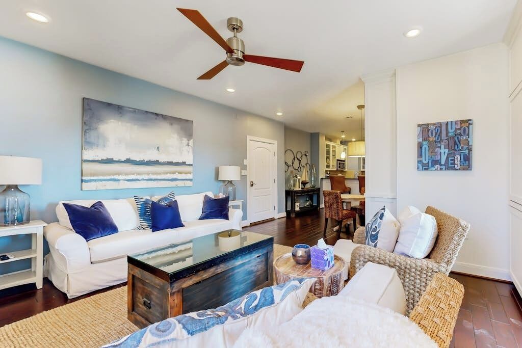A living room with a white sofa, a coffee table, a wicker chair, a ceiling fan, and artwork on the walls.