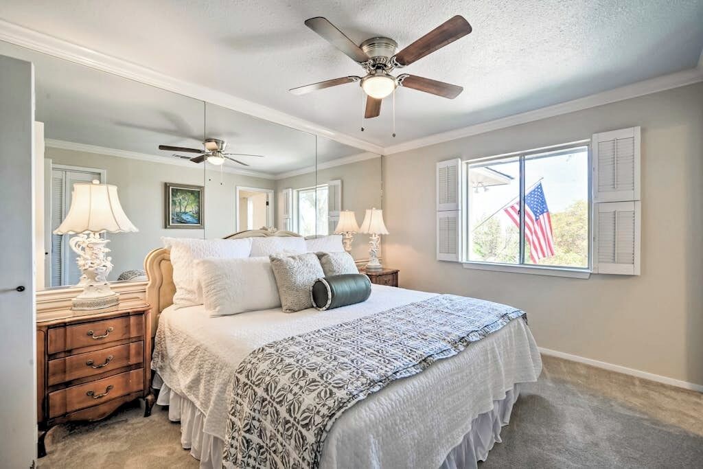 Une chambre à coucher avec un lit, des tables de chevet, des lampes, un ventilateur de plafond et une fenêtre ornée d’un drapeau américain.