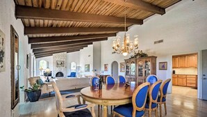 Uma ampla área de jantar com uma mesa de madeira e cadeiras estofadas azuis, um lustre e uma cozinha ao fundo.
