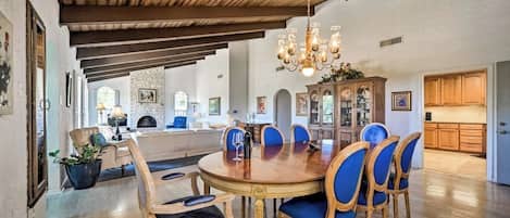 A spacious dining area with a wooden table and blue upholstered chairs, a chandelier, and a kitchen in the background.
