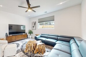 Una sala de estar moderna con un sofá modular, un televisor de pantalla plana, un ventilador de techo y un ventanal grande.