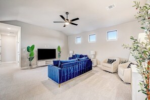 A spacious living room with a large flat-screen TV, a blue sofa, two armchairs, a ceiling fan, and a small window. - Exquisite Dog-friendly Home With Balcony, Enclosed Yard, Central AC, & W/D (Leander)