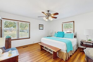 Ein Schlafzimmer mit einem Bett, einem Deckenventilator, einem Fenster mit Jalousien, einem Nachttisch und einer Bank.