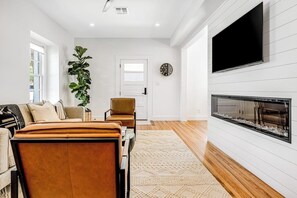 A modern living room with a fireplace, a large TV, and a plant. - Historic Home a Block From Main St With Hot Tub, Sauna, Fast Wifi & Firepit (Fredericksburg)