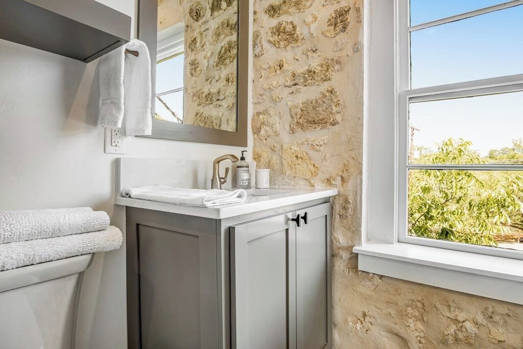 Un baño con pared de piedra, ventana con vistas, lavamanos con artículos de aseo y perchero para toallas.