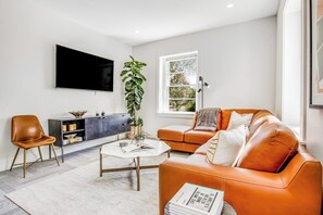 A modern living room with a sofa, a coffee table, a TV, and a plant. - Historic Home a Block From Main St With Hot Tub, Sauna, Fast Wifi & Firepit (Fredericksburg)