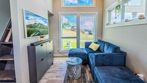 A modern living room with a blue sofa, a flat-screen TV, and a staircase. - Modern Tiny Home With Loft, Kitchen, Cozy Patio and Shared Pools, Great Location (Fredericksburg)