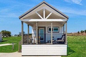 A small, single-story house with a porch, a door, and a window. - Modern Tiny Home With Loft, Kitchen, Cozy Patio and Shared Pools, Great Location (Fredericksburg)