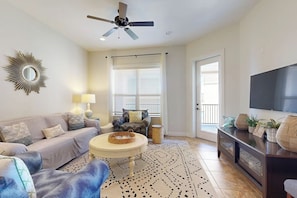 A living room with a sofa, a coffee table, a TV, and a ceiling fan. - Hill Country Corner Condo at The Waters - Central AC, Lake Views & Pool (Horseshoe Bay)
