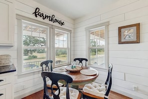A dining area with a round table, four chairs, and a view through large windows. - Cozy Ranch Home With Full Kitchen, Living Room, Hot Tub, Pet-friendly & Fireplace (Fredericksburg)