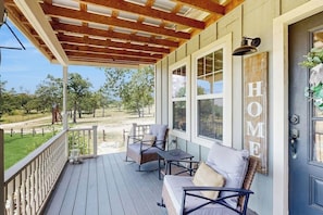 A covered porch with a wooden ceiling, a seating area with a sofa and chairs, large windows, and a 'HOME' sign on the door. - Cozy Ranch Home With Full Kitchen, Living Room, Hot Tub, Pet-friendly & Fireplace (Fredericksburg)